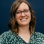 Smiling woman with shoulder-length hair and glasses, wearing a green patterned blouse, against a dark blue background.