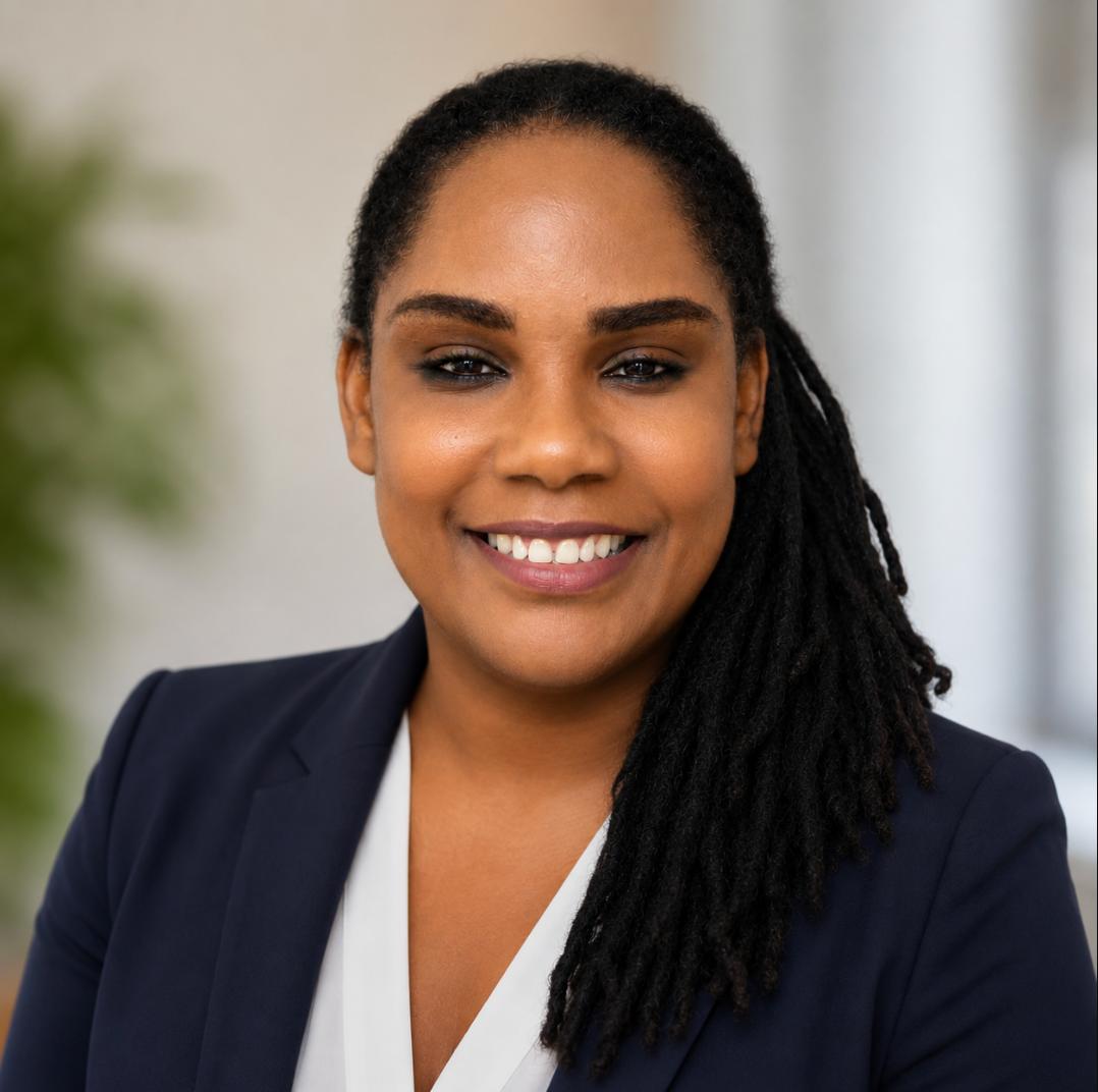 Professional headshot of a smiling woman with dark dreadlocks, wearing a navy blazer and white blouse.
