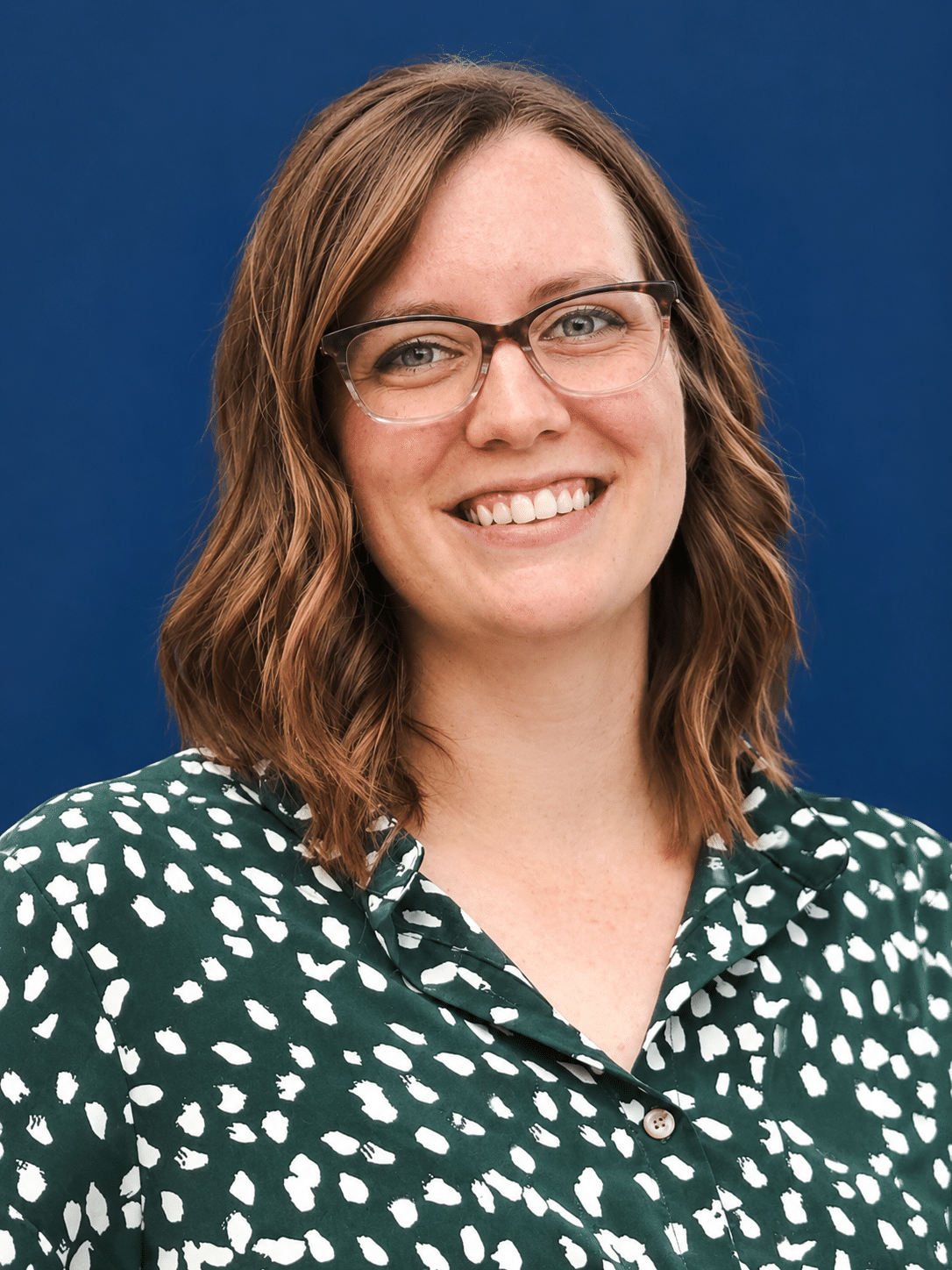 Smiling woman with glasses against a blue background, wearing a green patterned blouse.