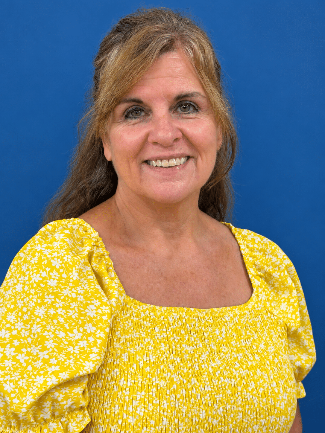Smiling woman with light brown hair in a yellow floral blouse against a blue background.